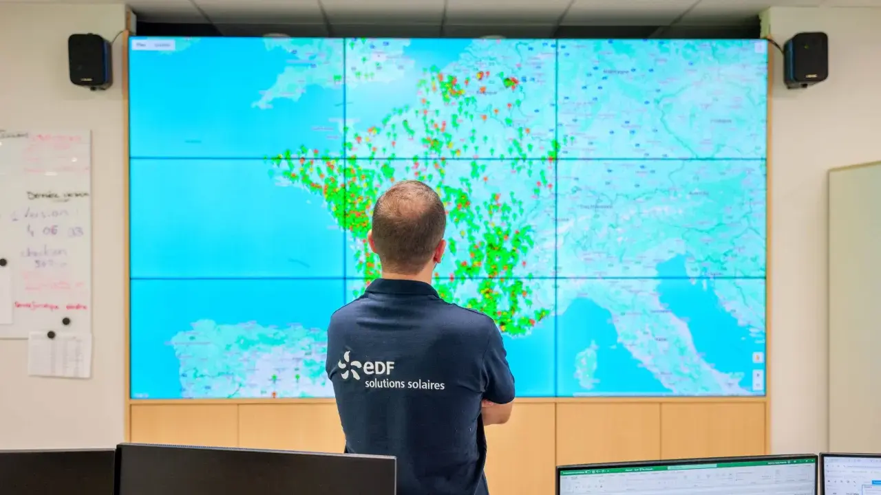 Un homme regarde une carte de la France sur un grand écran, avec des points verts et rouges. Il porte un polo "EDF solutions solaires", peut-être pour surveiller les installations et éviter une arnaque panneau solaire.