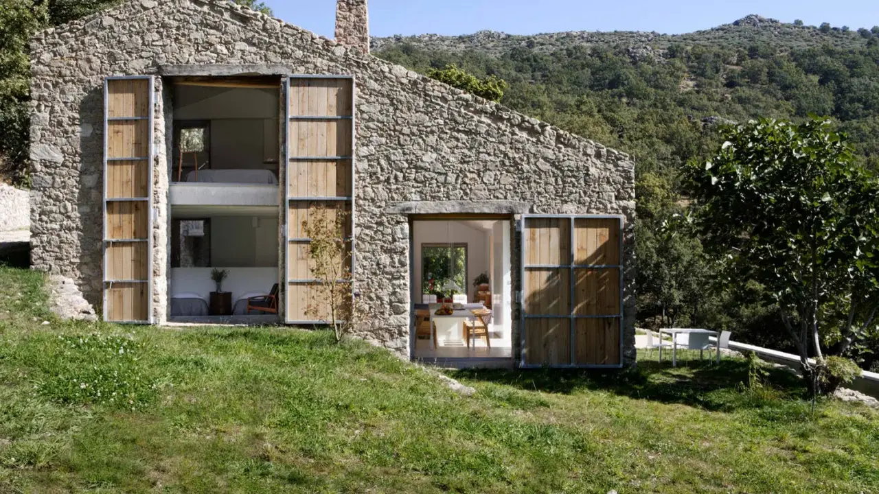 Una casa in pietra con grandi porte in legno, perfetta per chi ama vivere in campagna. Si intravedono interni moderni e un paesaggio collinare rigoglioso.