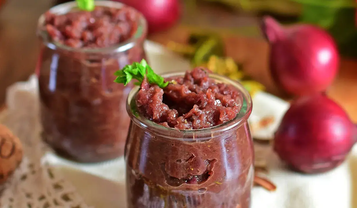 Dwa słoiczki z domowym, aromatycznym chutney z czerwonej cebuli, ozdobione listkiem zieleniny.