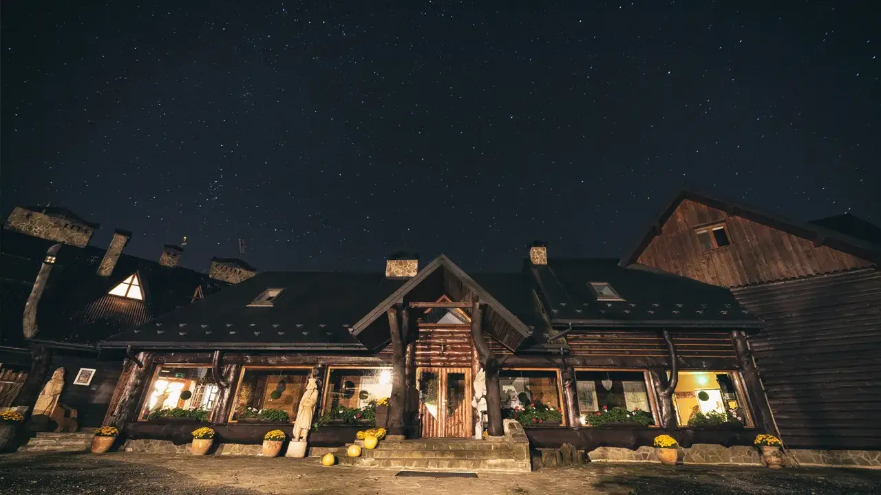 Nocne niebo nad bacówką Biały Jeleń. Drewniany budynek z oknami oświetlonymi ciepłym światłem, otoczony donicami z kwiatami.