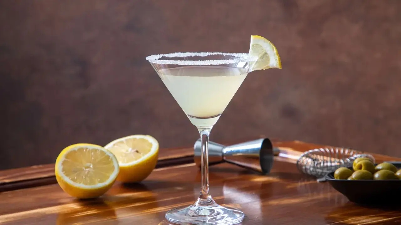 Orzeźwiający drink z martini, ozdobiony plasterkiem cytryny i solnym rantem. Obok leżą połówki cytryn, oliwki i akcesoria barmańskie. Idealny na domowe drinki z martini.
