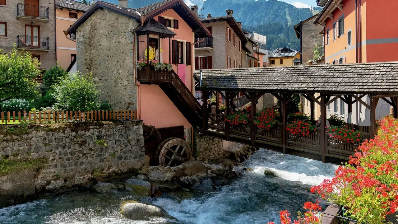 Un pittoresco ponte di legno attraversa un torrente impetuoso, fiancheggiato da case colorate e fiori rossi. Cosa vedere in questo incantevole borgo alpino.