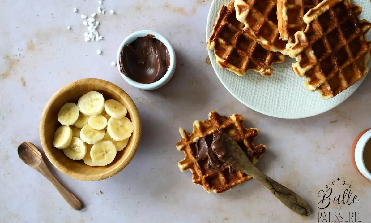 Une gaufre bubble est garnie de pâte à tartiner au chocolat. À côté, des rondelles de banane et une cuillère en bois.