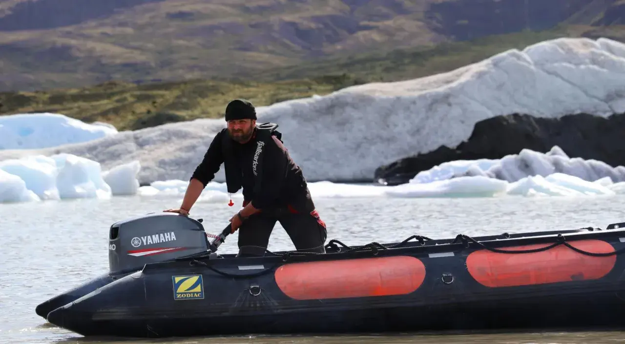 Un homme tente de démarrer un moteur 2 temps sur un Zodiac, mais il cale. Derrière lui, des icebergs flottent dans un lac glaciaire.