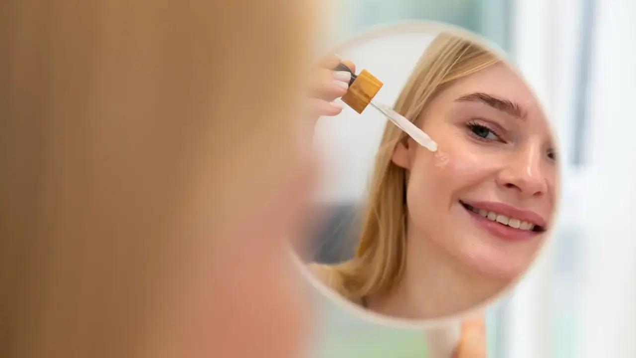 Mujer rubia sonríe mientras se aplica aceite facial con gotero en la mejilla. Un paso esencial en su rutina de cuidado de la piel, ya sea aceite facial antes o después de la crema.