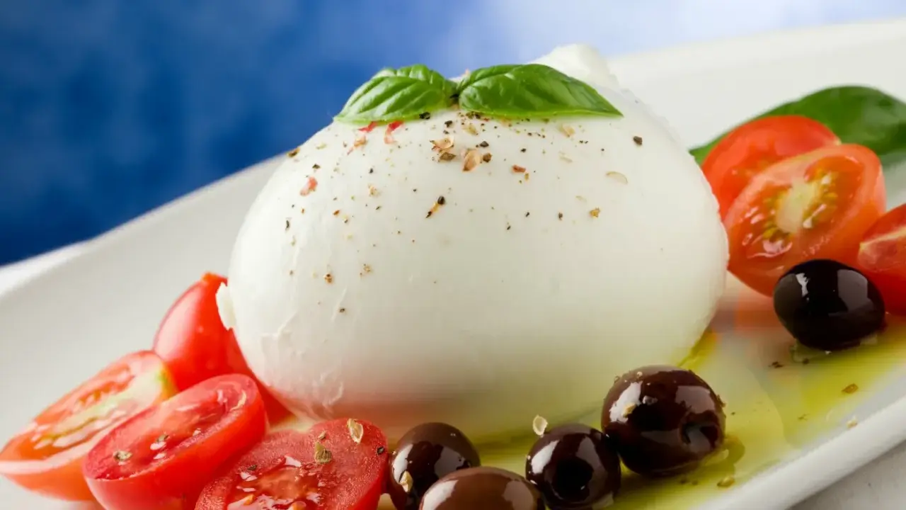 Duża kulka mozzarelli di bufala z pomidorkami koktajlowymi, oliwkami i bazylią na białym talerzu.