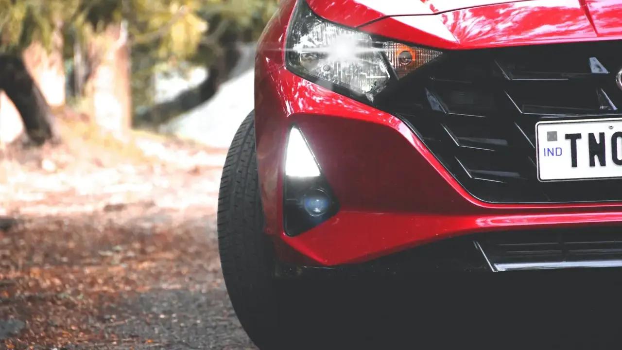 Czerwony Hyundai i20 z zapalonymi światłami dziennymi i reflektorami. Widać fragment maski, grill i tablicę rejestracyjną.