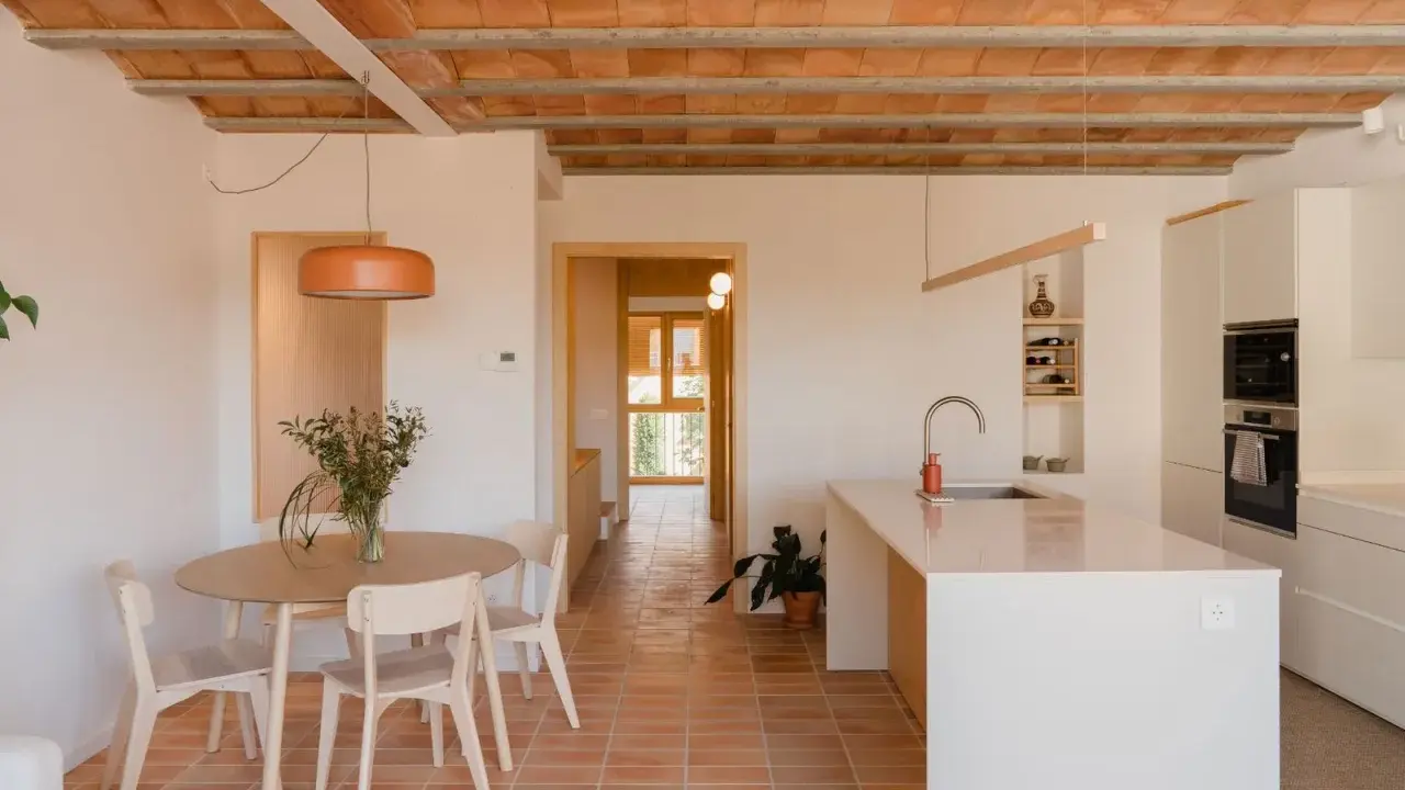 Comedor y cocina luminosos en una casa vieja decorada con poco dinero. Techos de vigas de madera y baldosas de terracota.