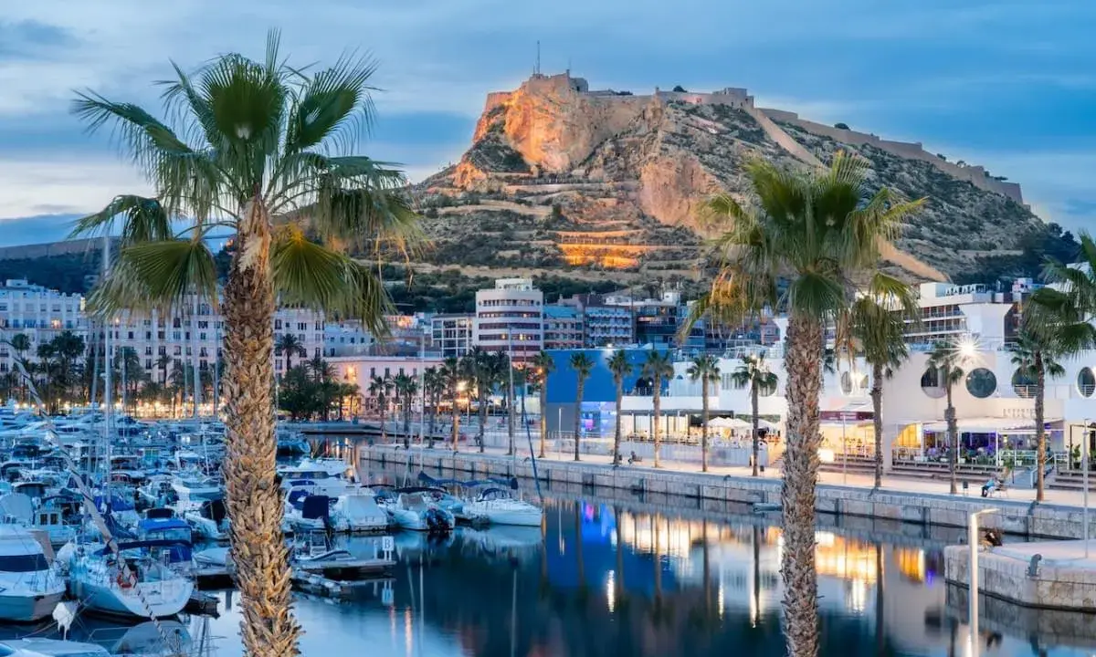 Palme e barche nel porto di Alicante, con il Castello di Santa Barbara illuminato sullo sfondo.