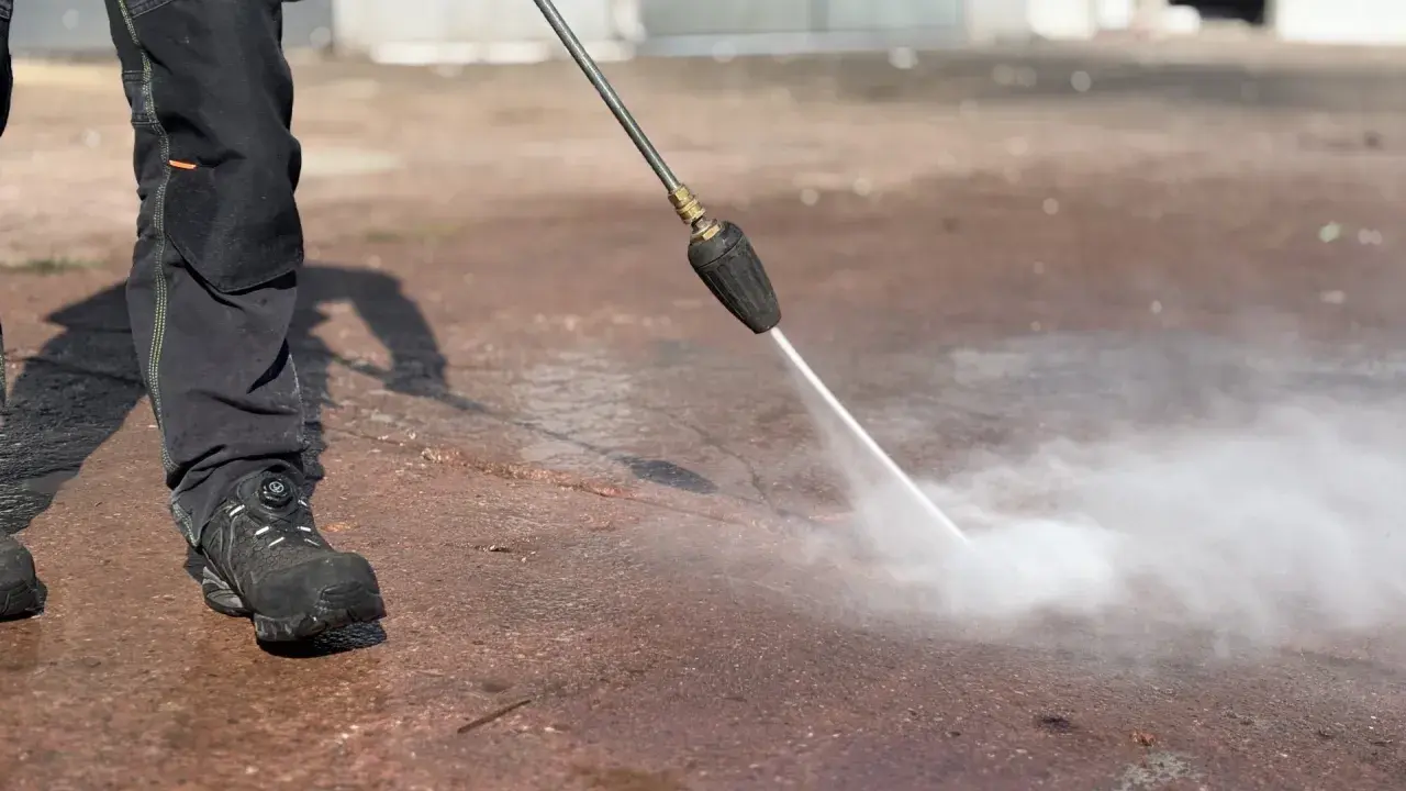 Osoba w roboczych spodniach i butach czyści mokrą nawierzchnię za pomocą myjki ciśnieniowej z dyszą rotacyjną.