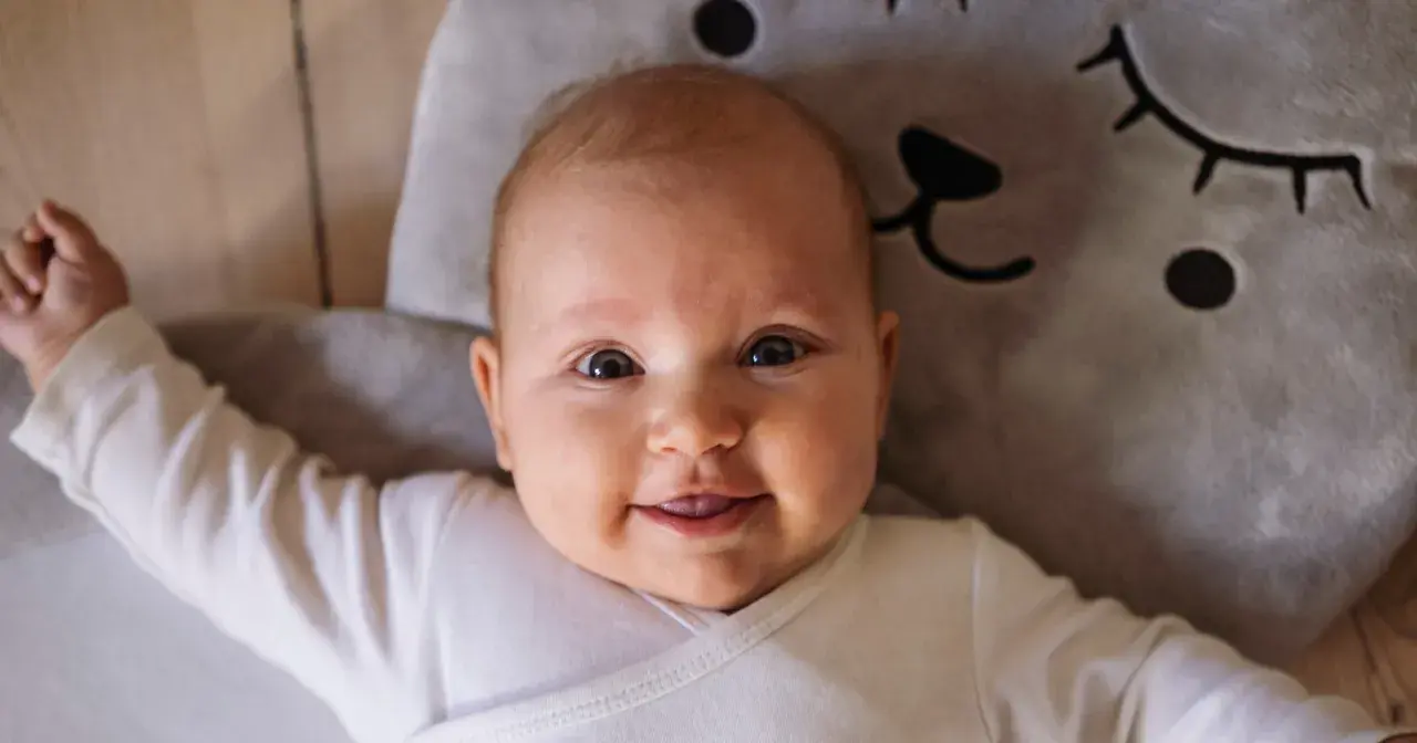Uśmiechnięte niemowlę w białym body leży na miękkiej poduszce w kształcie misia. To mądre imię dla chłopca, które przyniesie mu szczęście.