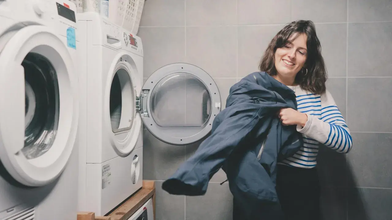 Kobieta z uśmiechem trzyma granatową kurtkę, przygotowując się do prania Gore-Tex. Obok stoi otwarta suszarka.