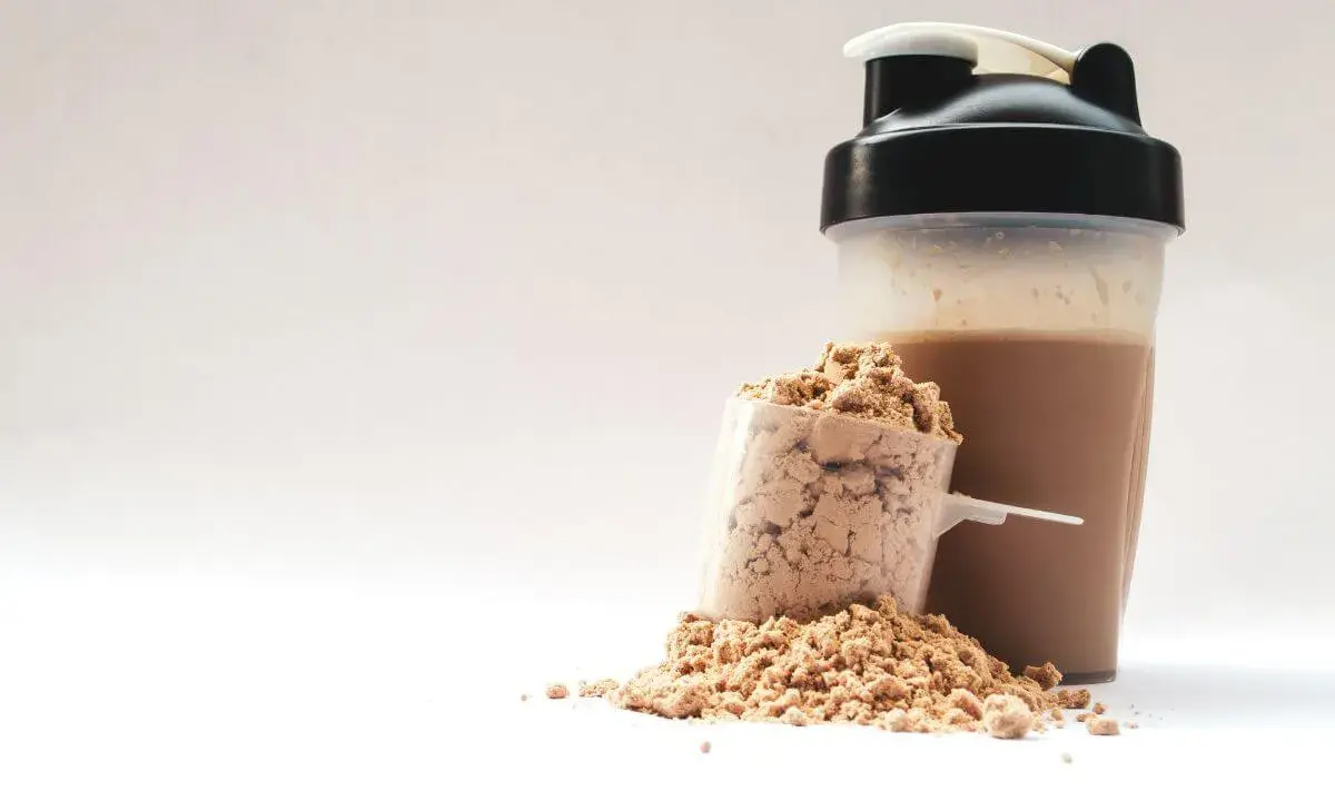 Ein Löffel Proteinpulver neben einem fertigen Proteinshake im Shaker. Perfekt für den proteinreichen Start in den Morgen.