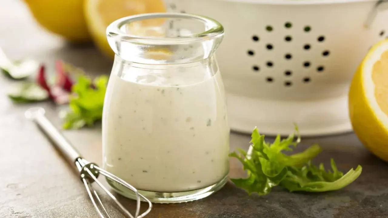 Ein Glas cremiges Ranch Dressing, daneben ein Schneebesen, Salatblätter und Zitronenspalten. Perfekt für dein Ranch Dressing Rezept!