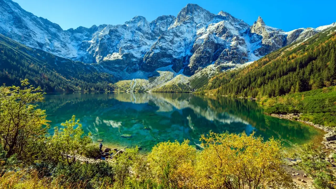 Widok na Morskie Oko z wysokości. Turkusowa tafla jeziora odbija ośnieżone szczyty Tatr. Jesienne drzewa otaczają brzeg.
