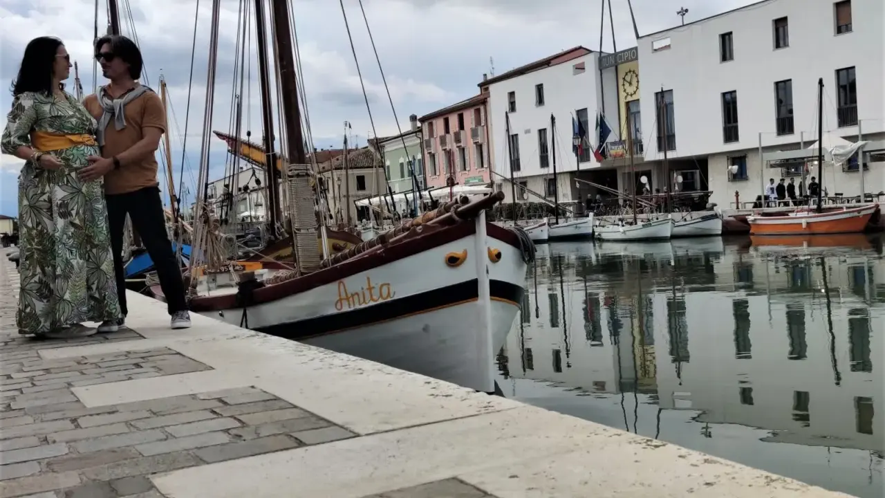 Coppia in attesa a Cesenatico, con barche d'epoca ormeggiate e il Municipio sullo sfondo. Un ricordo speciale prima di San Marino.