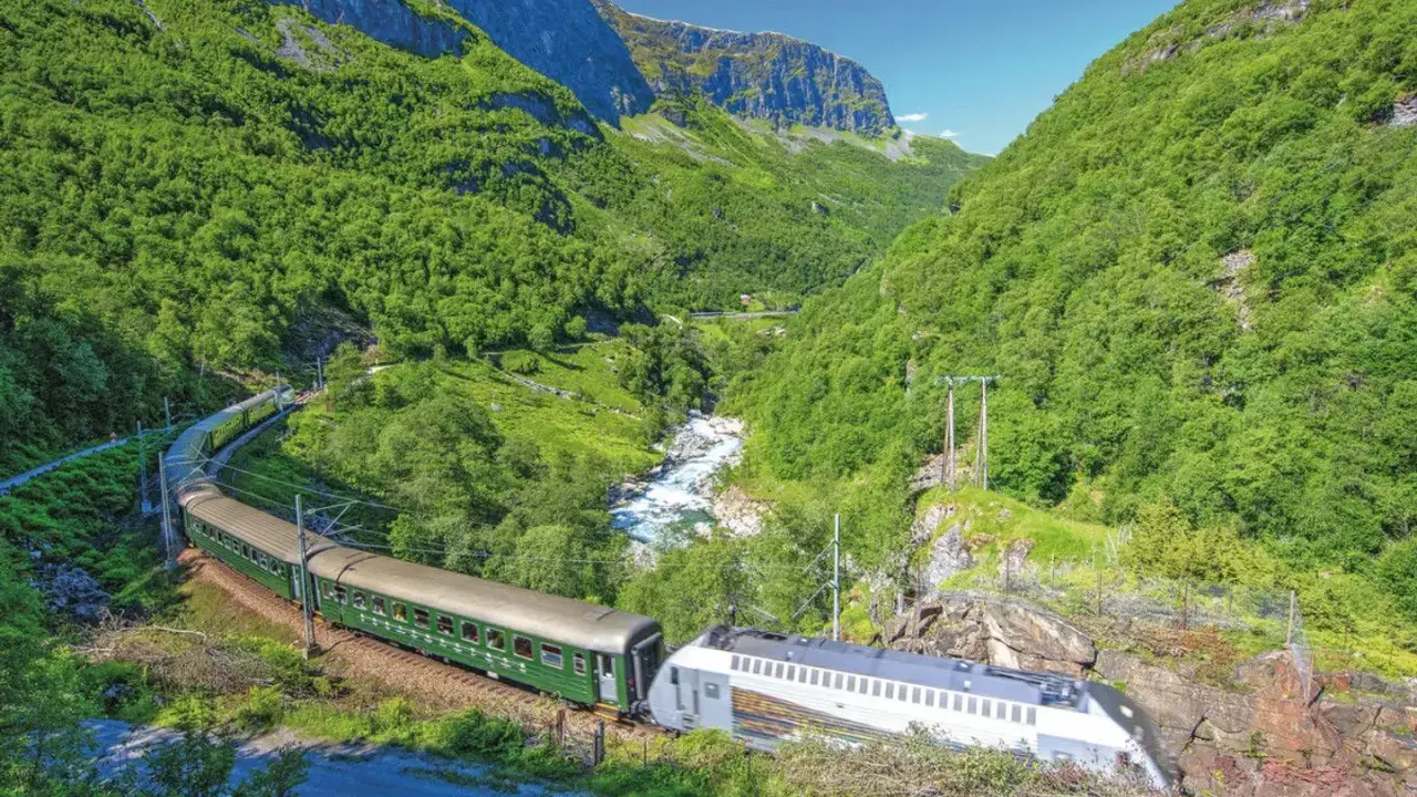 Grüner Zug fährt durch ein bergiges Tal mit Fluss. Die Fahrt von Bergen nach Oslo ist ein unvergessliches Erlebnis.