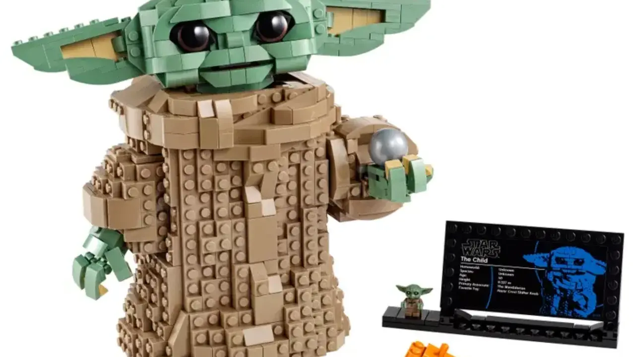 Figurka Grogu z klocków LEGO, znana z gier LEGO Star Wars, trzyma srebrną kulę. Obok stoi mała figurka i tabliczka informacyjna.