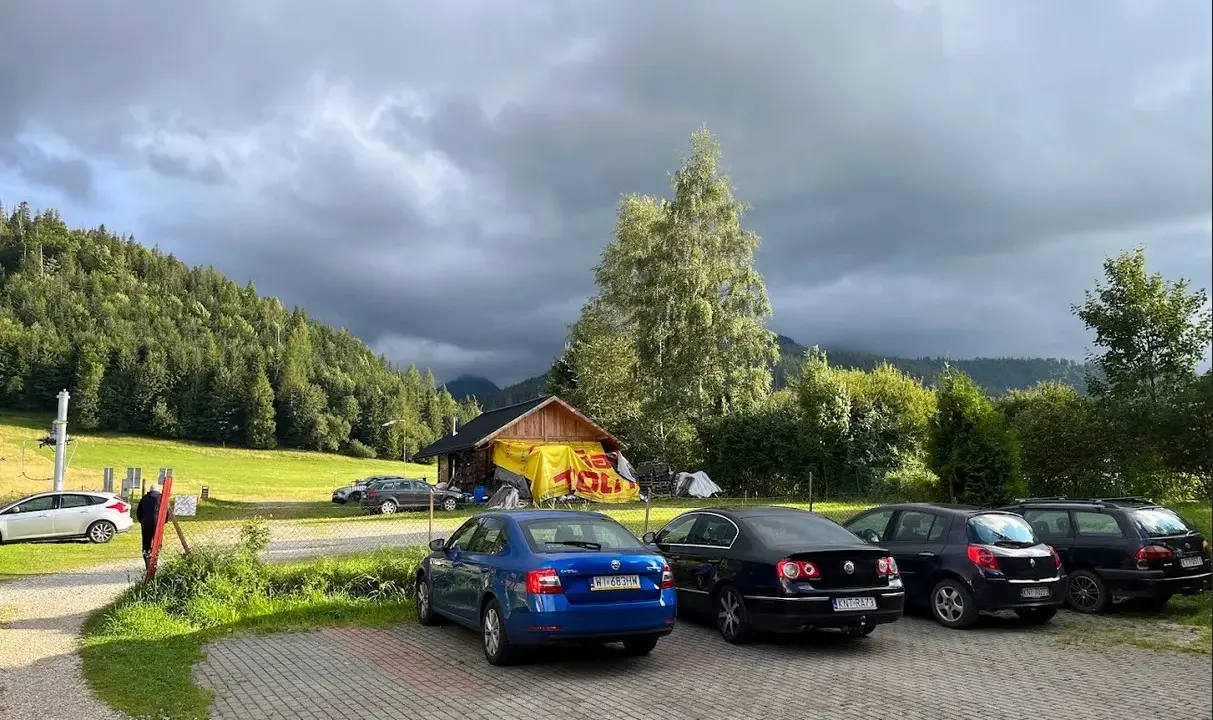 Kasprowy Wierch parking pod burzowym niebem. Kilka samochodów zaparkowanych przy górskiej chatce z żółtym banerem.
