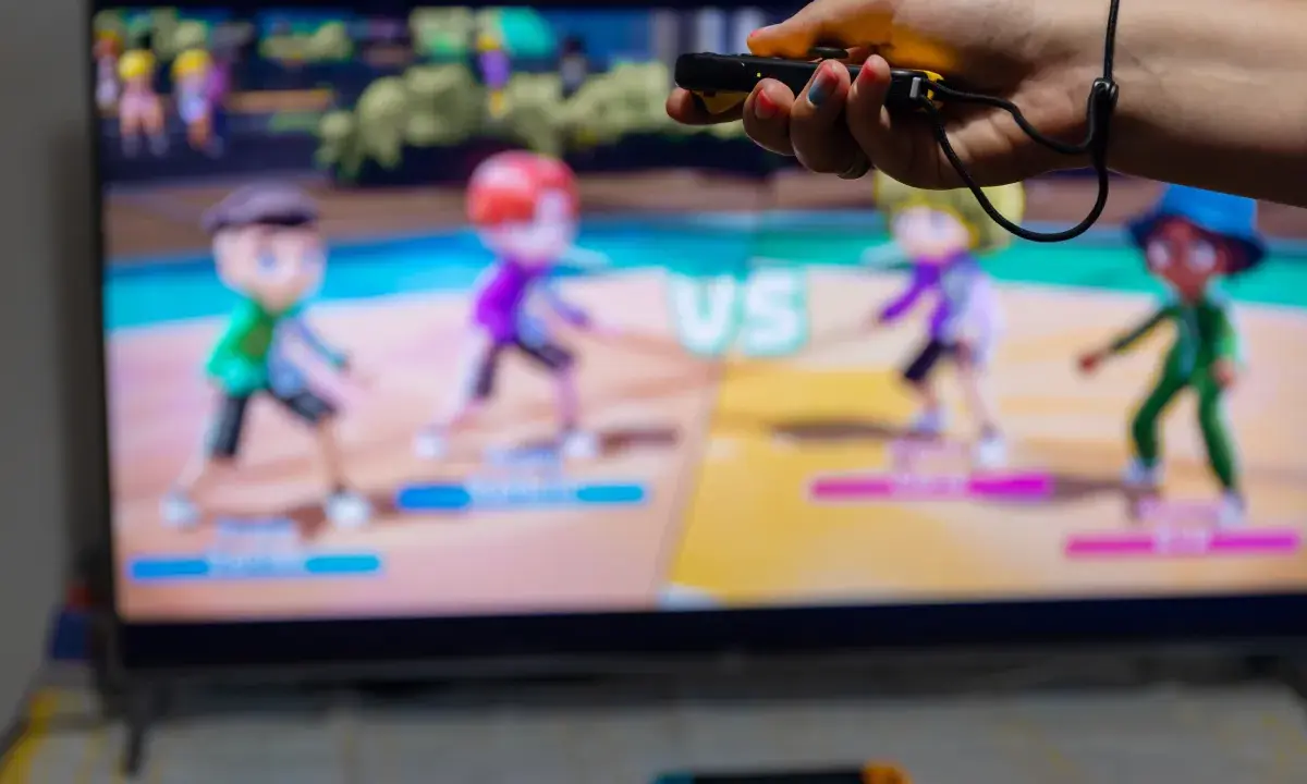 Ręka trzymająca kontroler do gry, na ekranie telewizora widać kolorowe postacie z gry ruchowej Nintendo Switch.