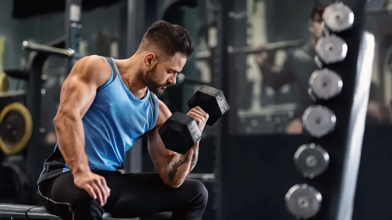 Ein Mann trainiert im Fitnessstudio mit Hanteln. Er fragt sich, wie oft ins Gym pro Woche er gehen sollte, um fit zu bleiben.