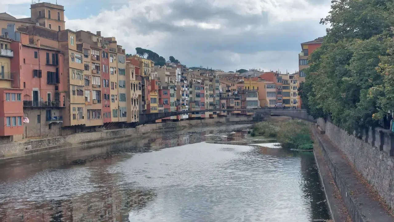 Bunte Häuserfronten säumen den Fluss Onyar in der malerischen **Girona Altstadt**. Eine Brücke verbindet die Ufer, während sich die Wolken am Himmel spiegeln.