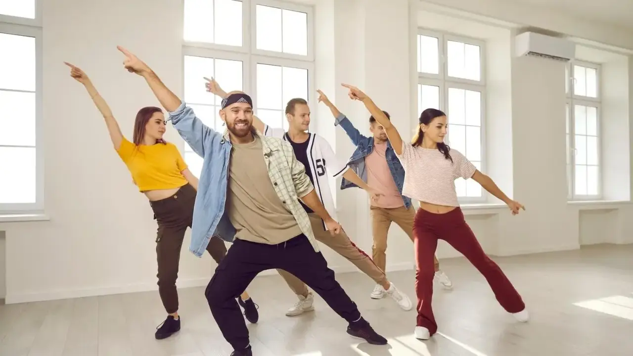 Grupa ludzi ćwiczy energiczne samba kroki w jasnej sali tanecznej, z rękami uniesionymi w rytm muzyki.