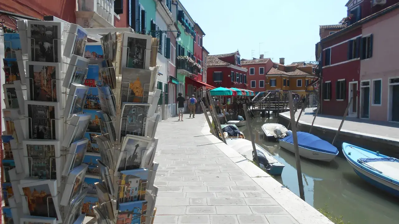 Cartoline di Murano, Burano e Torcello in vendita lungo un canale con case colorate e barche ormeggiate.