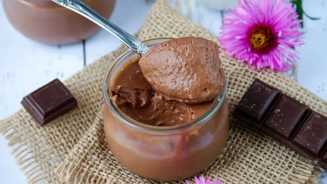 Une cuillère de crème au chocolat sans sucre, onctueuse et aérienne, sur un pot en verre. Des carrés de chocolat et des fleurs roses décorent la scène.