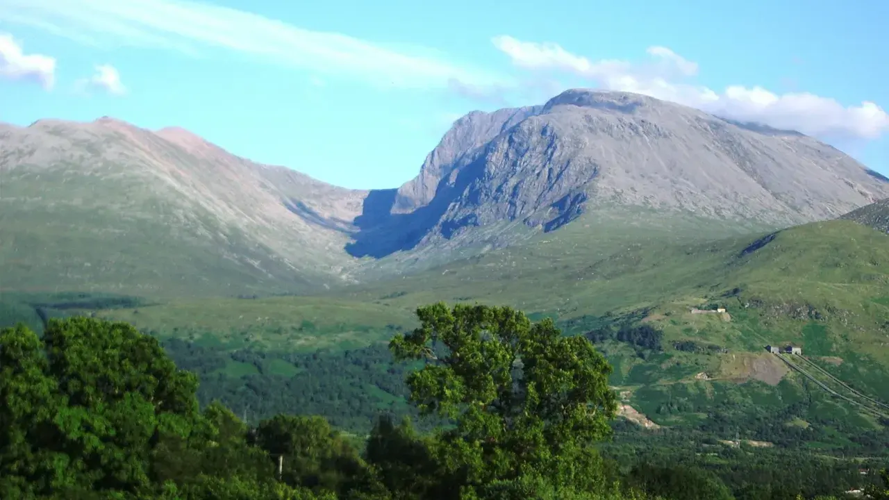Majestatyczny Ben Nevis wznosi się na tle błękitnego nieba z delikatnymi chmurami. Zielone zbocza górskie i bujna roślinność tworzą malowniczy krajobraz.