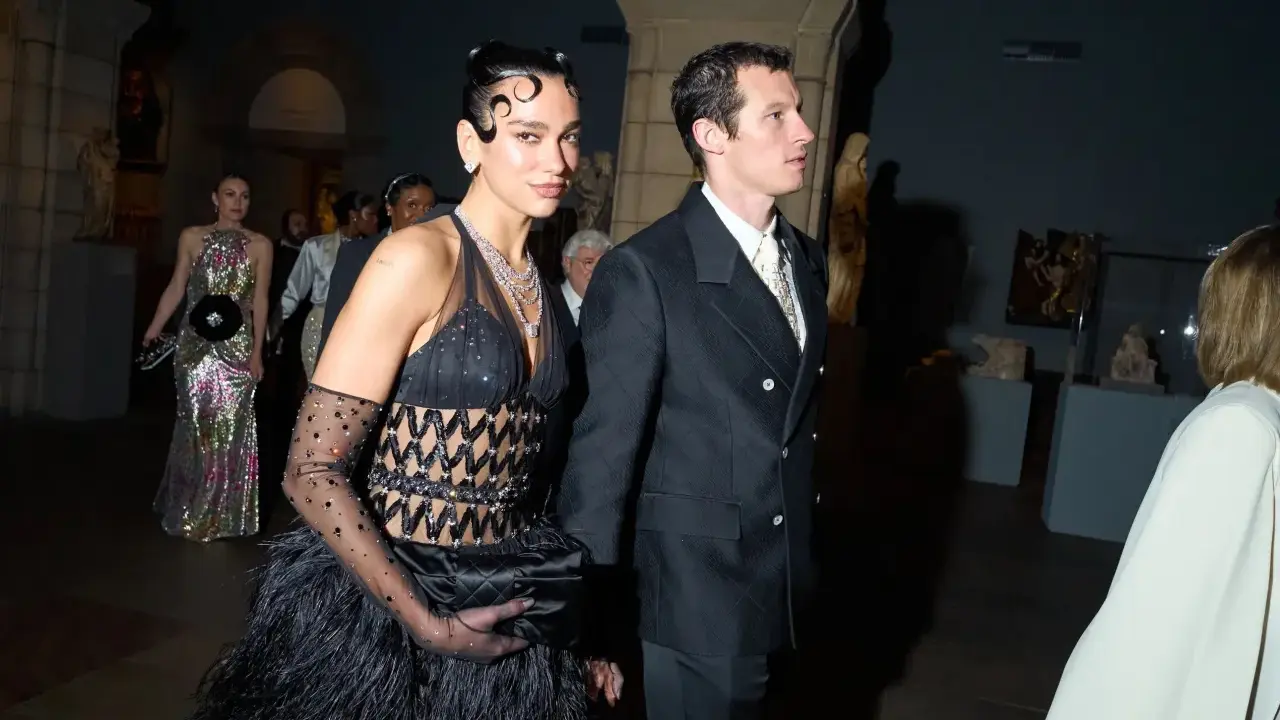 Dua Lipa stuns at the Met Gala in a black feathered gown and diamond necklace, walking with a companion.