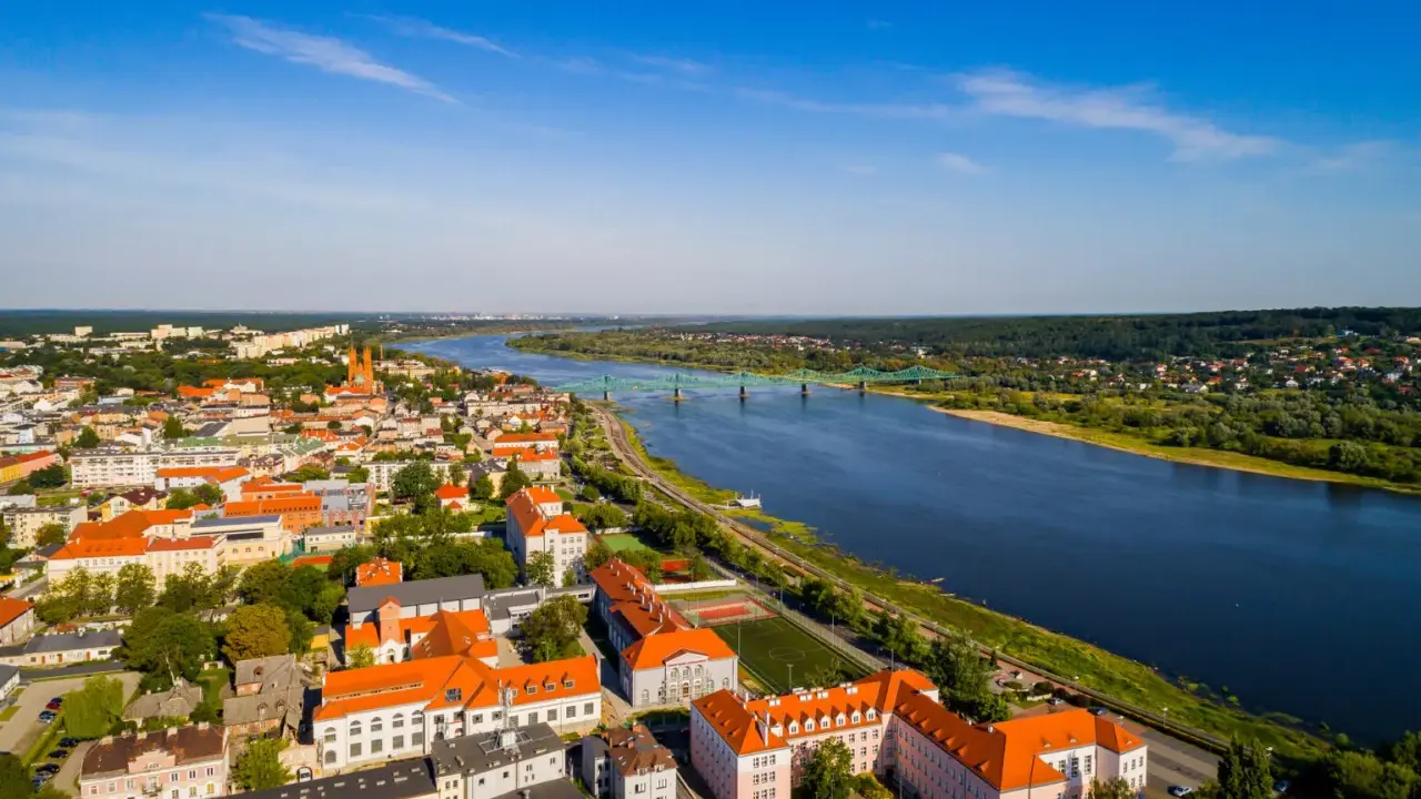 Widok z lotu ptaka na miasto nad Wisłą, z zielonym mostem i zabudowaniami w słoneczny dzień.