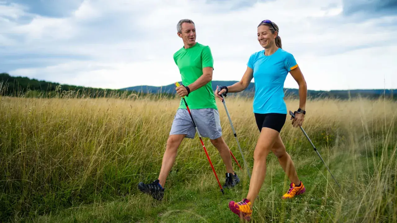 Ein Paar genießt das **power walking** in einer sonnigen Landschaft mit hohem Gras und bewaldeten Hügeln im Hintergrund.