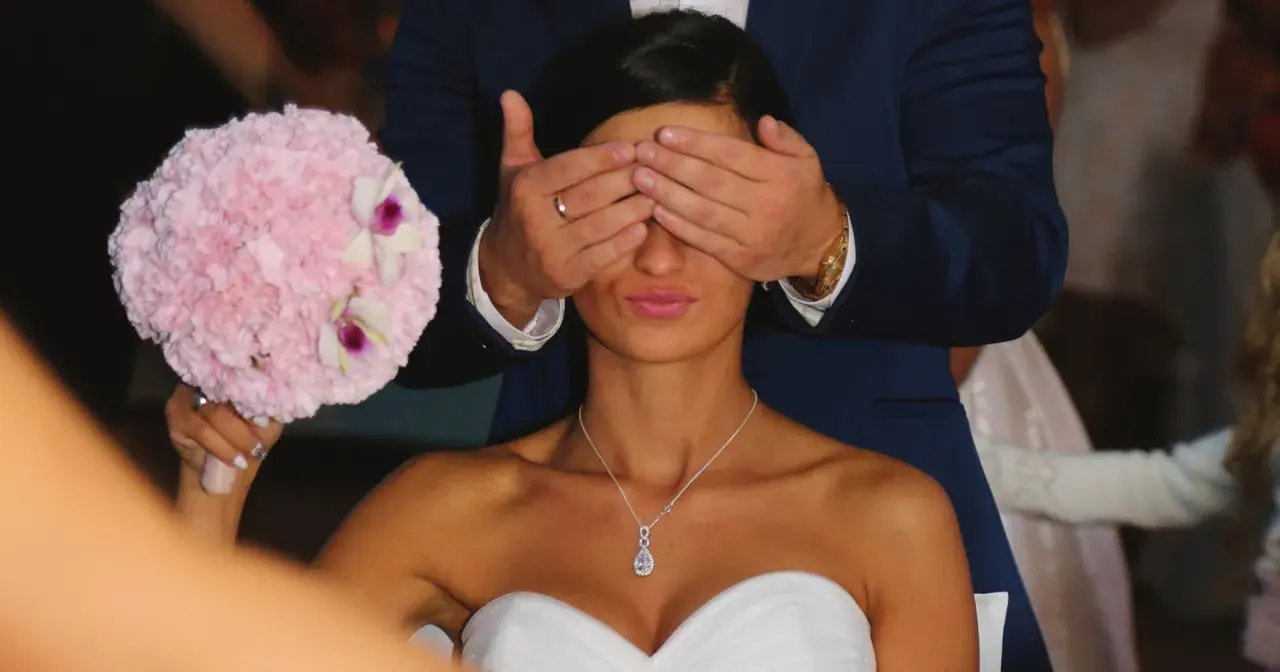 Pan młody zasłania oczy pannie młodej, która trzyma bukiet. To tradycja oczepin, co to za zwyczaj?