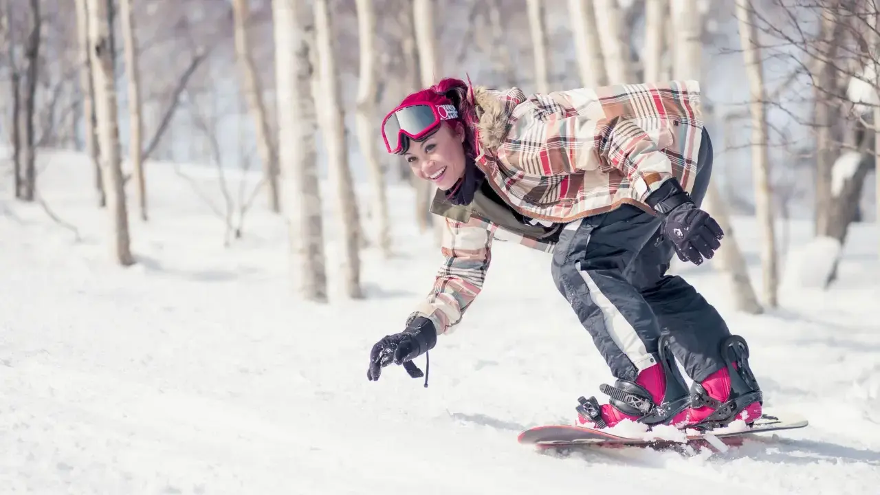 Uśmiechnięta snowboardzistka z różowymi włosami zjeżdża po ośnieżonym stoku wśród drzew. To świetna zabawa podczas chodzenia po górach zimą.
