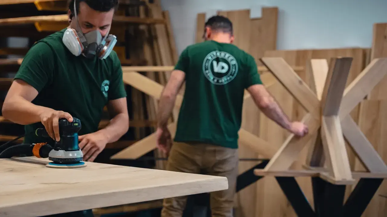 Un artisan porte un masque et utilise une ponceuse pour poncer une table en bois. Un autre artisan travaille sur un pied de table en bois.