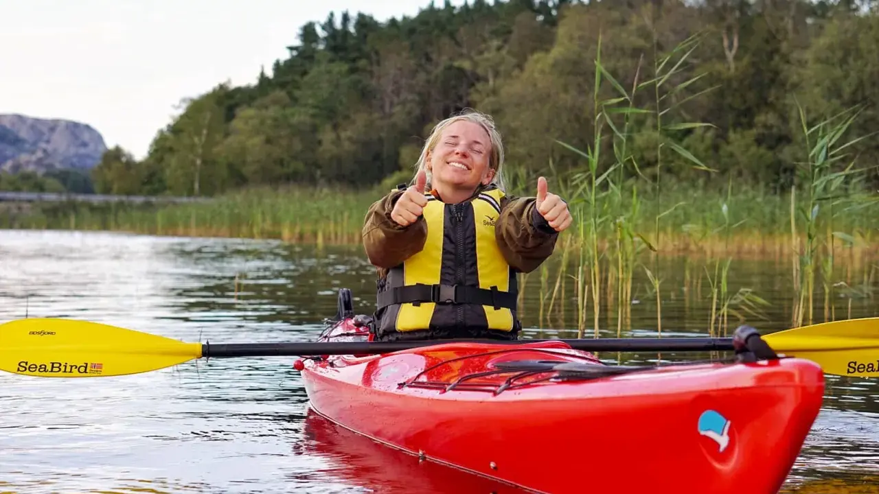 Glad kvinna i kajak med all kajak utrustning, ger tummen upp. Vacker natur med vass och träd i bakgrunden.