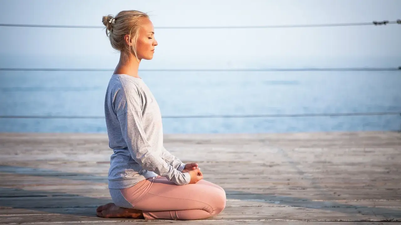 Kvinna med slutna ögon sitter i meditationsställning på en träbrygga vid havet, utövar progressiv avslappning.