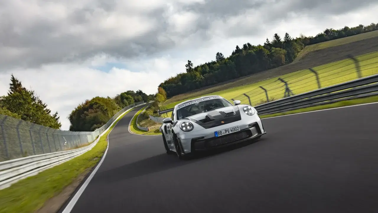 Biały Porsche 911 GT3 RS z czarnymi akcentami pędzi po torze wyścigowym wśród zielonych wzgórz i chmur.