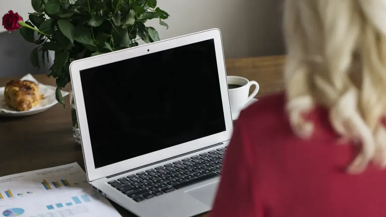 Un ordinateur portable ouvert sur un bureau en bois, avec une tasse de café, des roses et un croissant. Le branchement PC est prêt pour le travail.