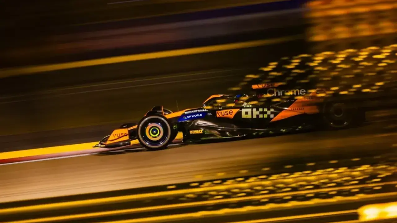 Un coche de **fórmula 1** naranja y negro acelera en la noche, dejando estelas de luz dorada.