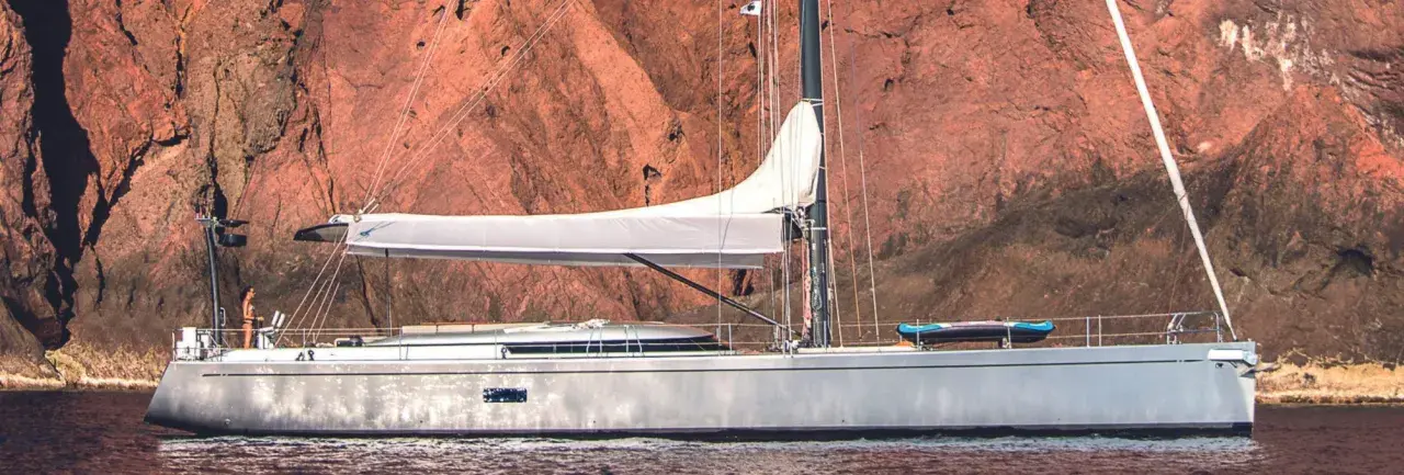 Un voilier Swan 76, avec une voile blanche déployée, navigue près d'une falaise rouge. Un kayak est rangé sur le pont.