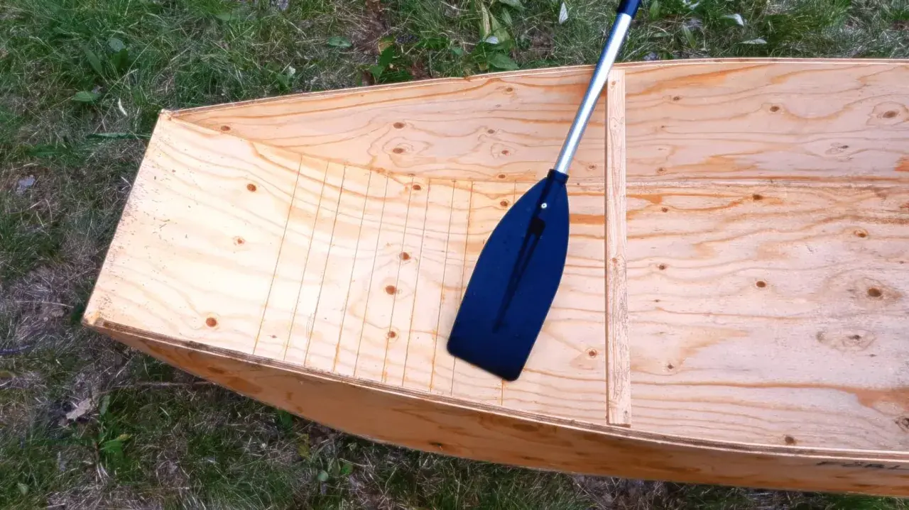 Bateau en contreplaqué brut, prêt pour la résine époxy. Une pagaie noire repose à l'intérieur.