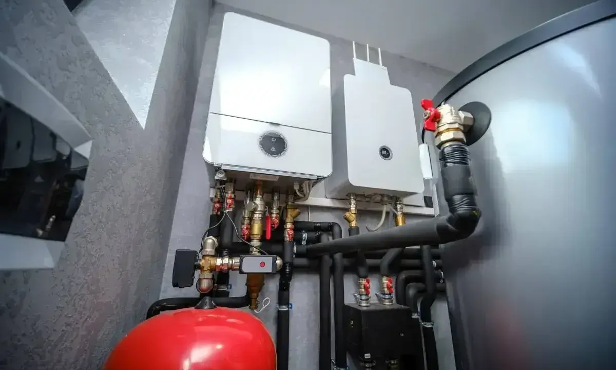 Dwa nowoczesne kotły gazowe wiszą na ścianie obok dużego, srebrnego bojlera. Pod nimi widoczny jest czerwony zbiornik przeponowy i skomplikowana instalacja hydrauliczna.