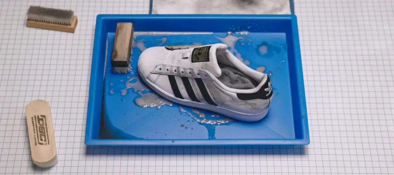 Ein schmutziger weißer Adidas Superstar Schuh liegt in einer blauen Wanne mit Wasser und Seife. Reinigungswerkzeuge liegen daneben.