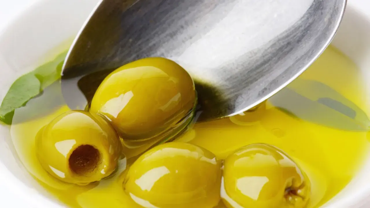 Kilka zielonych oliwek w złocistej oliwie, łyżka zanurzona w płynie. Oliwa z oliwek kcal - zdrowie i smak.