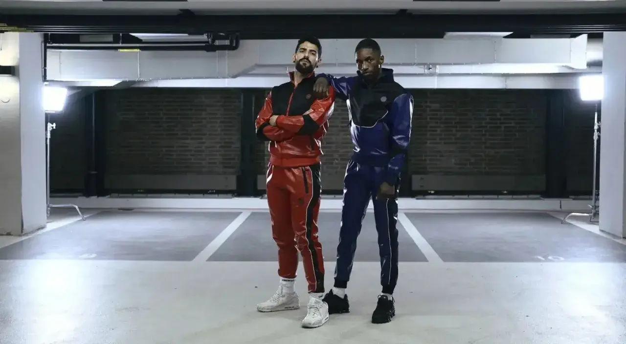 Two gay men in matching red and blue sportswear pose in a parking garage.