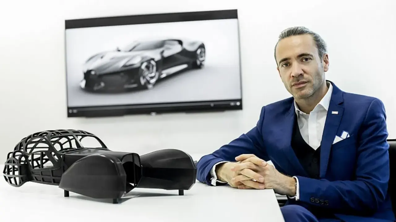 Ein Mann in einem blauen Anzug sitzt vor einem Bildschirm mit dem Bugatti La Voiture Noire. Daneben ein Modell des Autos.