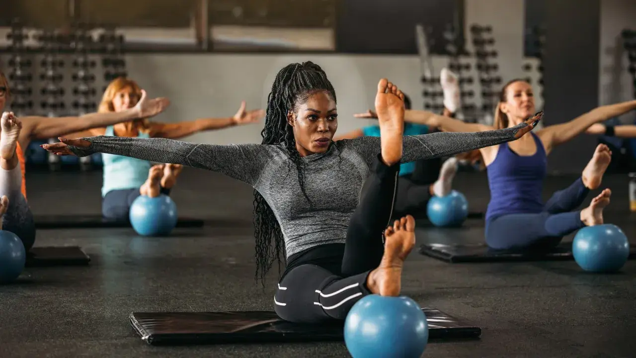 Frauen in einem Kurs, der Pilates Krafttraining macht, sitzen auf Matten mit kleinen blauen Bällen unter ihren Füßen.