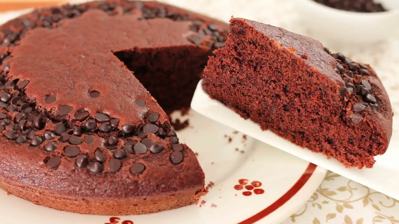 Una fetta di torta al cioccolato morbida, cosparsa di gocce di cioccolato, viene servita da una tortiera intera.